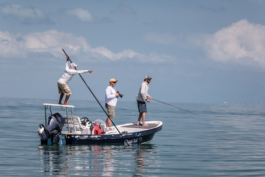 Three men on a flats boat fly fishing, one poling, one casting a fly rod, and one photographing them against a calm sea and cloudy sky