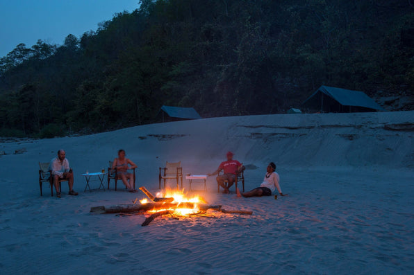 Travelers gather around a campfire on a sandy riverbank beside tents at dusk
