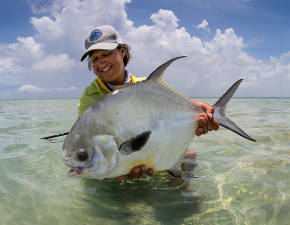 Fly fisher holding a large silver fish in shallow turquoise water under a cloudy sky