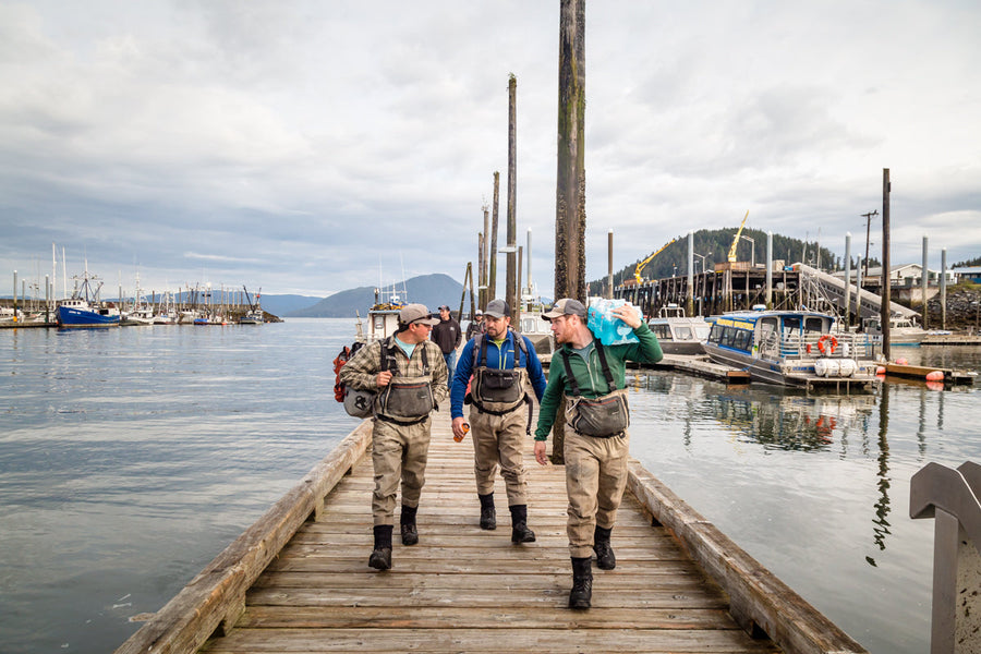 Three fly-fishing anglers in waders walking along a wooden dock carrying gear toward boats at an overcast marina