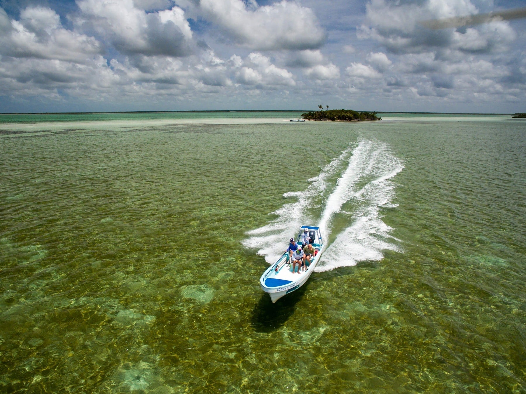 Small skiff with three people speeding across clear shallow green water toward a palm-fringed islet under cloudy sky
