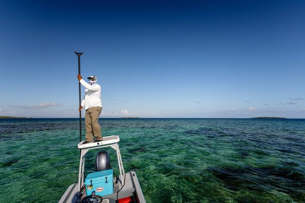 Fly fisher in white sun-protective clothing stands on a flats boat and uses a push pole in shallow turquoise water