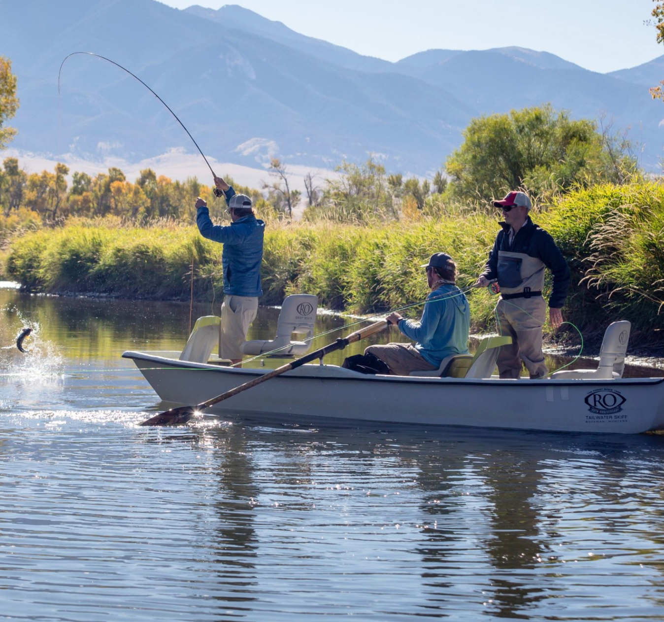 Three fly fishers casting from a boat on a Montana river with mountains and reeds in the background
