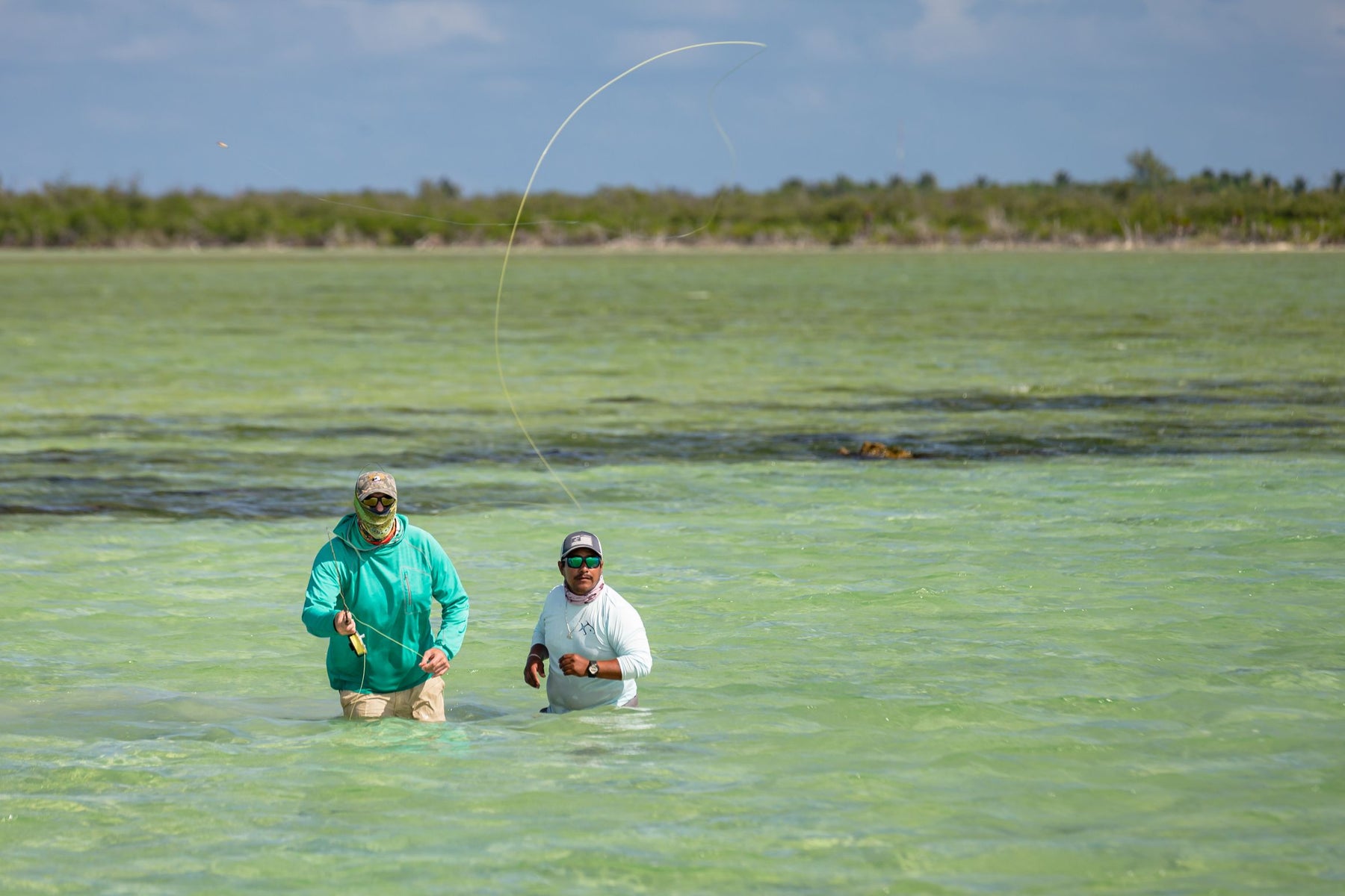 Two anglers fly fishing in shallow turquoise water with a shoreline behind them