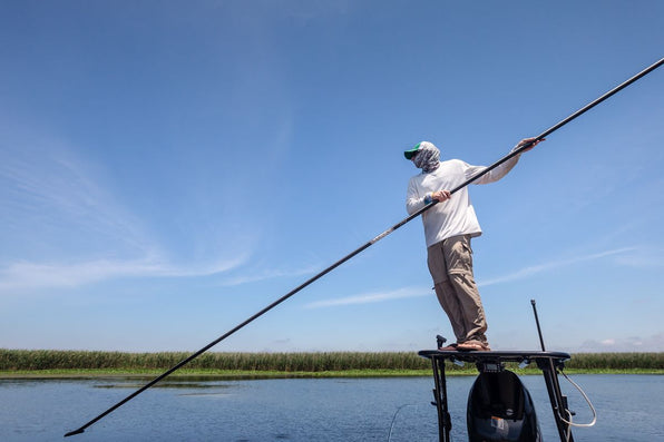 Fly fisher poles a shallow-water boat through Louisiana marsh grass under a clear blue sky