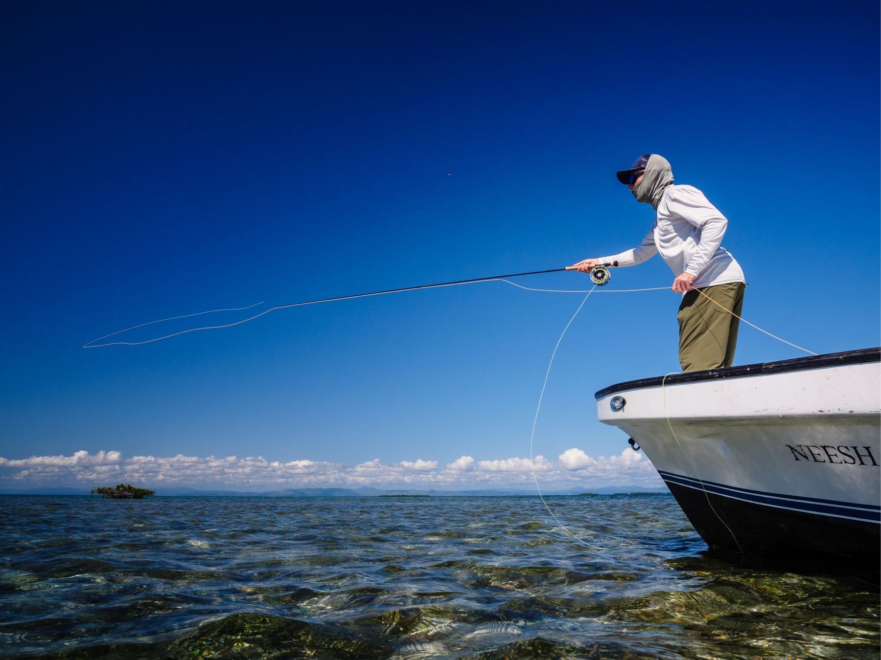 Angler fly fishing from a boat over shallow ocean water beneath a clear blue sky
