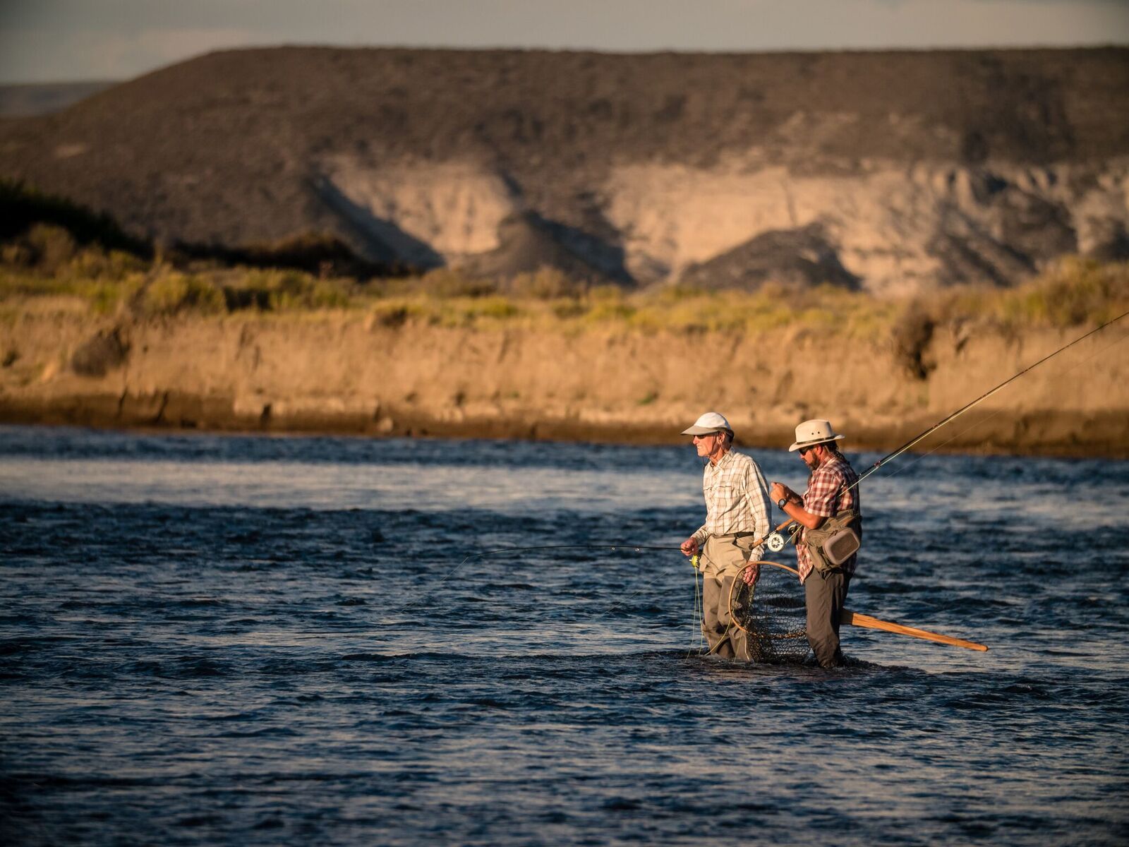 Two anglers fly fishing in a river against a rugged Patagonian landscape