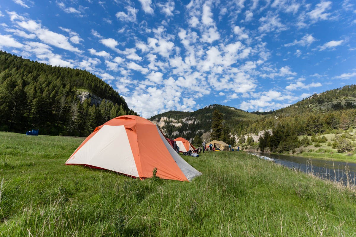 Campers set up orange tents beside a river in a forested mountain valley
