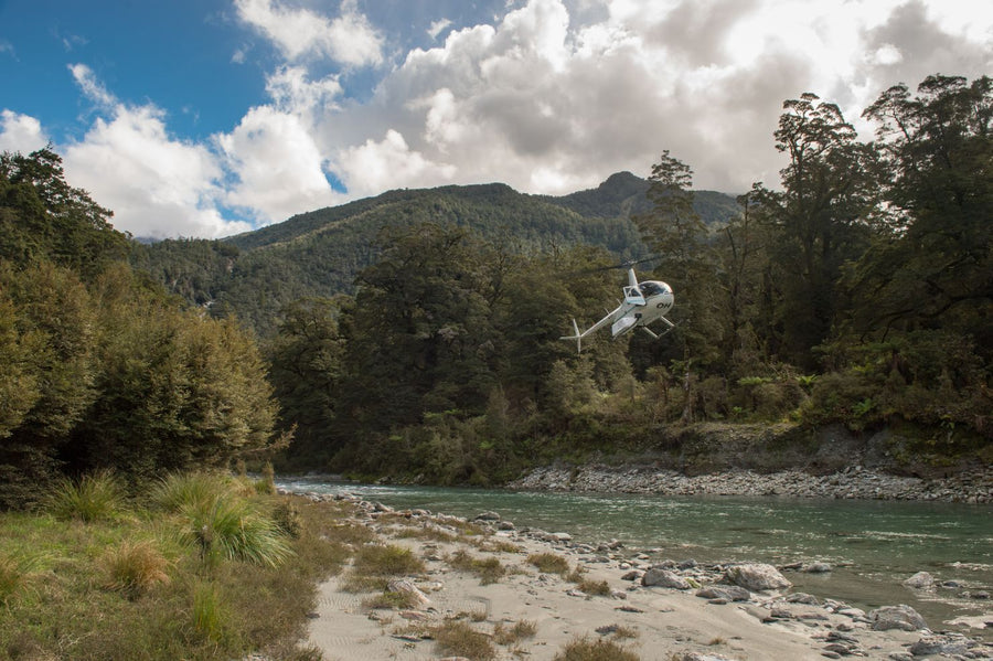 Helicopter hovering above a clear river in New Zealand during a fly fishing trip, forested hills and sandy riverbank