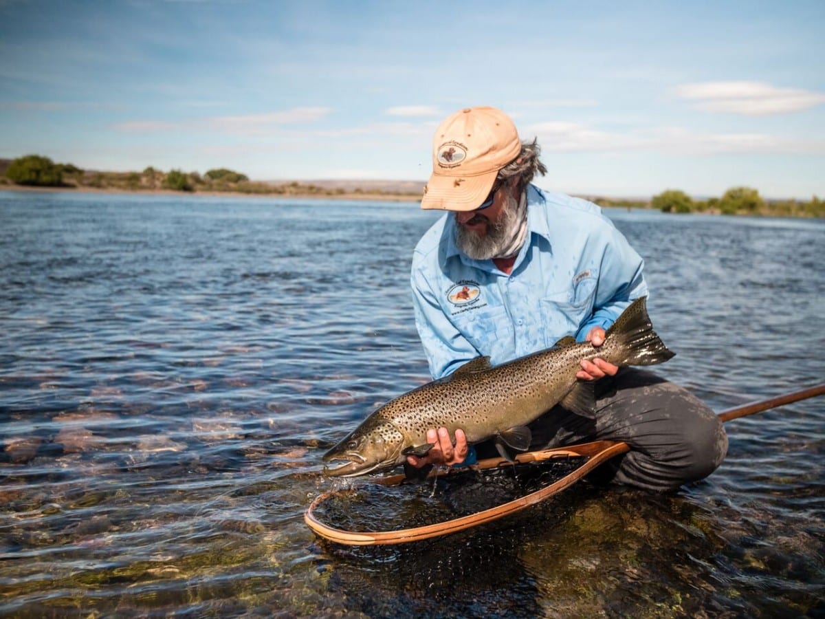 yellow-dog-flyfish Fly angler kneeling in a shallow river holding a large brown trout above a wooden landing net with river and sky behind