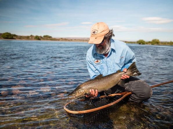 yellow-dog-flyfish Fly angler kneeling in a shallow river holding a large brown trout above a wooden landing net with river and sky behind