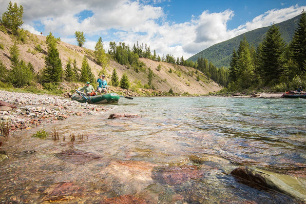 Fly fishers navigate a green raft on a river through a forested mountain landscape