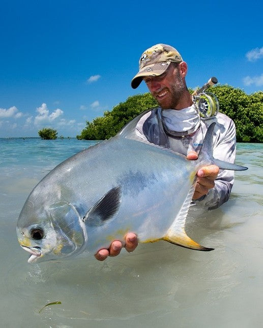 Fly angler holding a silver permit in shallow tropical flats with a fly rod over his shoulder and mangroves in the background