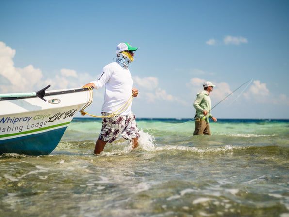 Guide wading and pulling a skiff while an angler casts a fly rod in shallow turquoise Belize waters on a sunny day