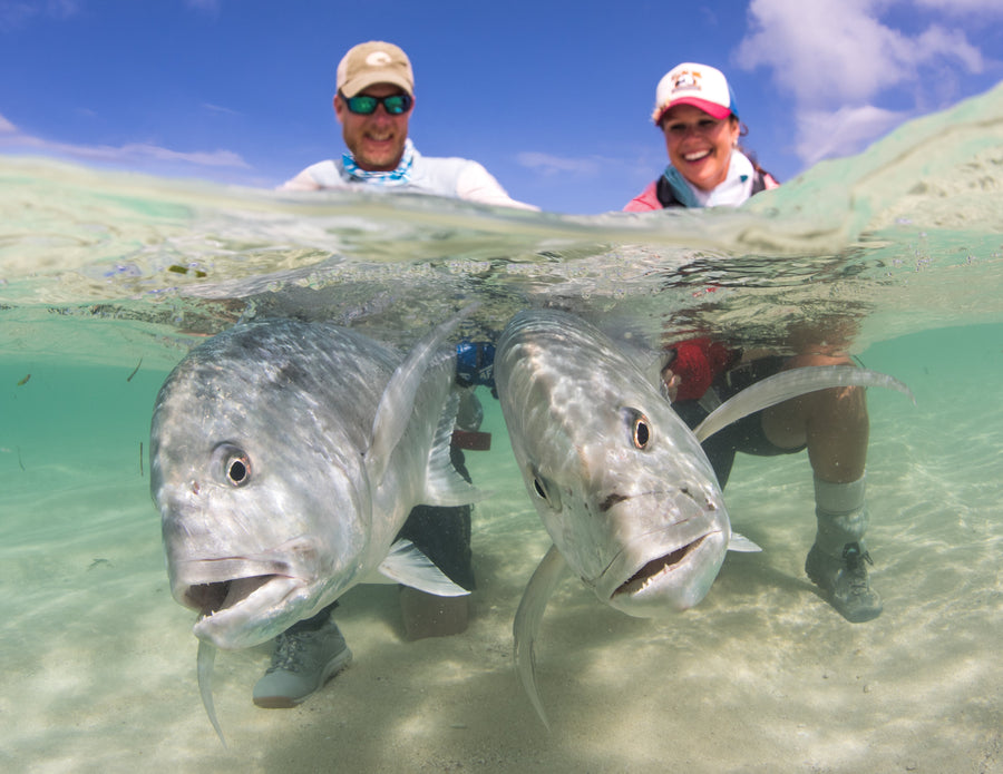 Two anglers knee-deep in clear turquoise water holding two large trevally near the camera at Providence Atoll