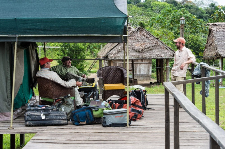 Three anglers talking on a wooden camp deck with fly fishing gear piled nearby and thatched jungle huts behind them