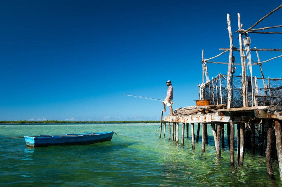 Man fly fishing from a rustic wooden pier over shallow turquoise Cuban waters with a small blue skiff nearby