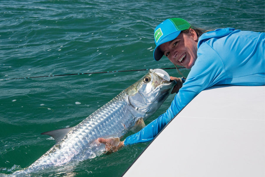 Female angler smiling from a boat while releasing a large tarpon into green water during a fly fishing trip