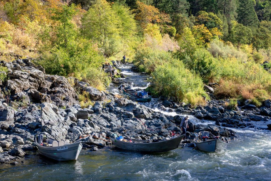 Three drift boats on a rocky Rogue River shore during a Fly Fishing Trips outing, guides loading gear, wooded banks