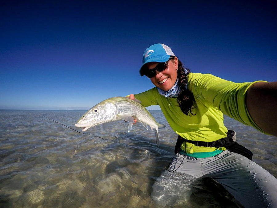 Angler smiling while holding a bonefish and wading a shallow sand flat under a clear blue sky on a fly fishing trip