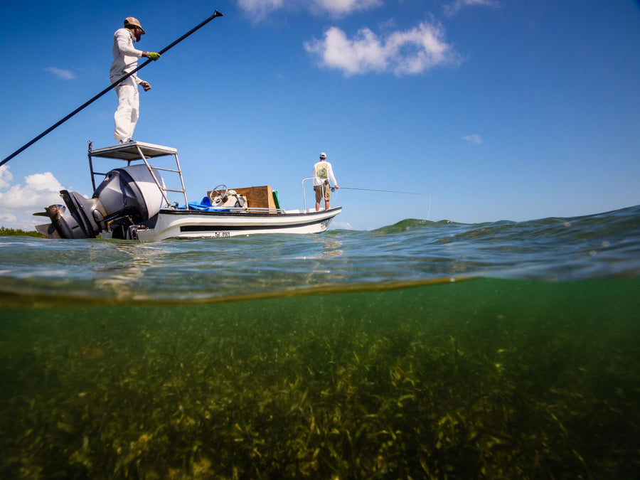Two anglers fly fishing from a flats skiff, one poling from a raised platform and one casting from the bow over clear shallow seagrass