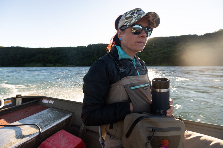 Woman angler in waders and sunglasses standing in a drift boat holding a travel mug, lake and forested shoreline behind her