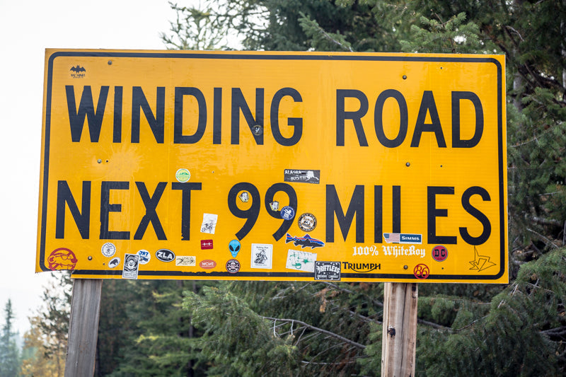 Yellow road sign reading WINDING ROAD NEXT 99 MILES at a forested roadside with stickers on the sign