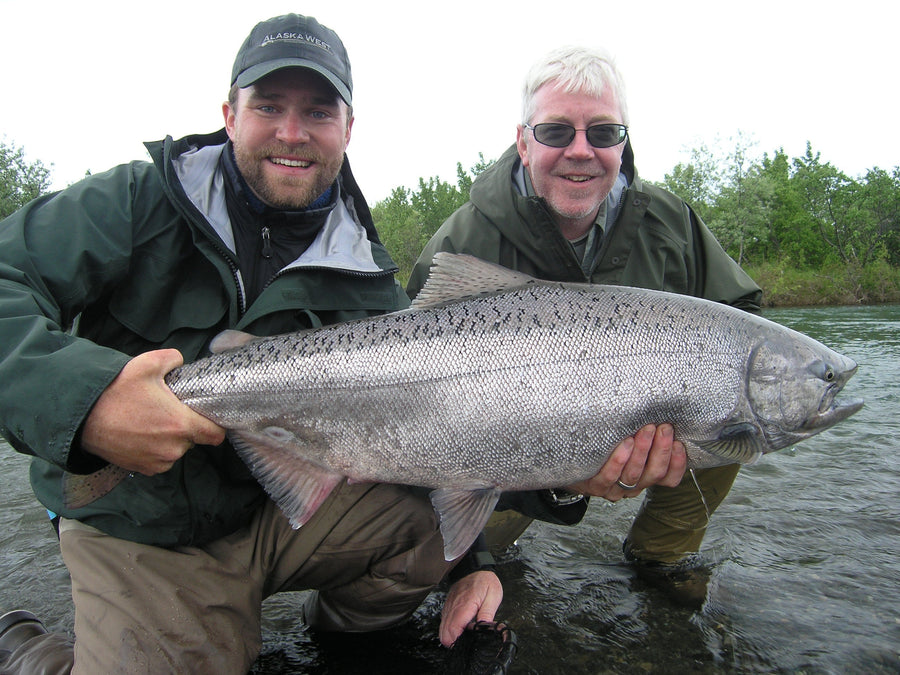 Two anglers smiling while holding a large silver salmon in a river on an Alaska West fly fishing trip