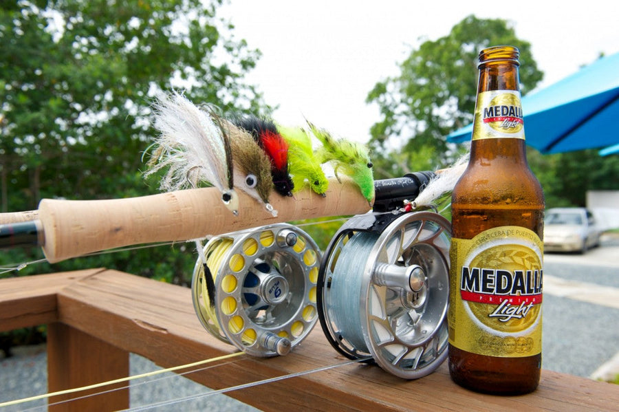 Fly rod with two reels and three colorful tied flies resting on a wooden railing next to a Medalla Light beer bottle outdoors