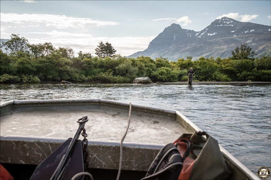 View from a drift boat of an angler wading and fly fishing in a river with a mountain backdrop
