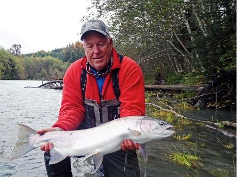 Angler holding a large steelhead while standing in a river with forested banks during a fly fishing trip