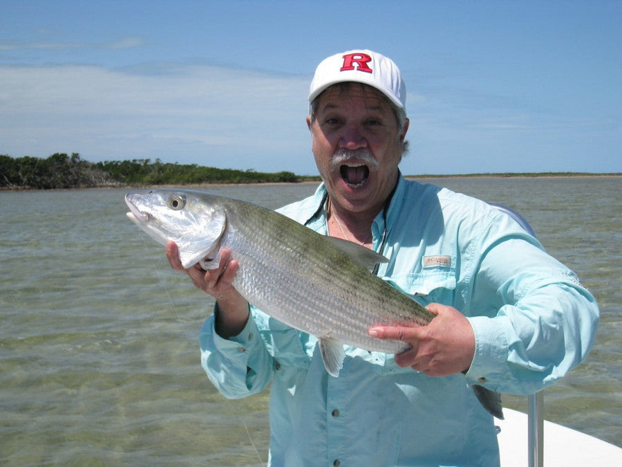 Angler in a light blue shirt and white cap holding a large bonefish on a flats skiff in shallow Bahamian waters