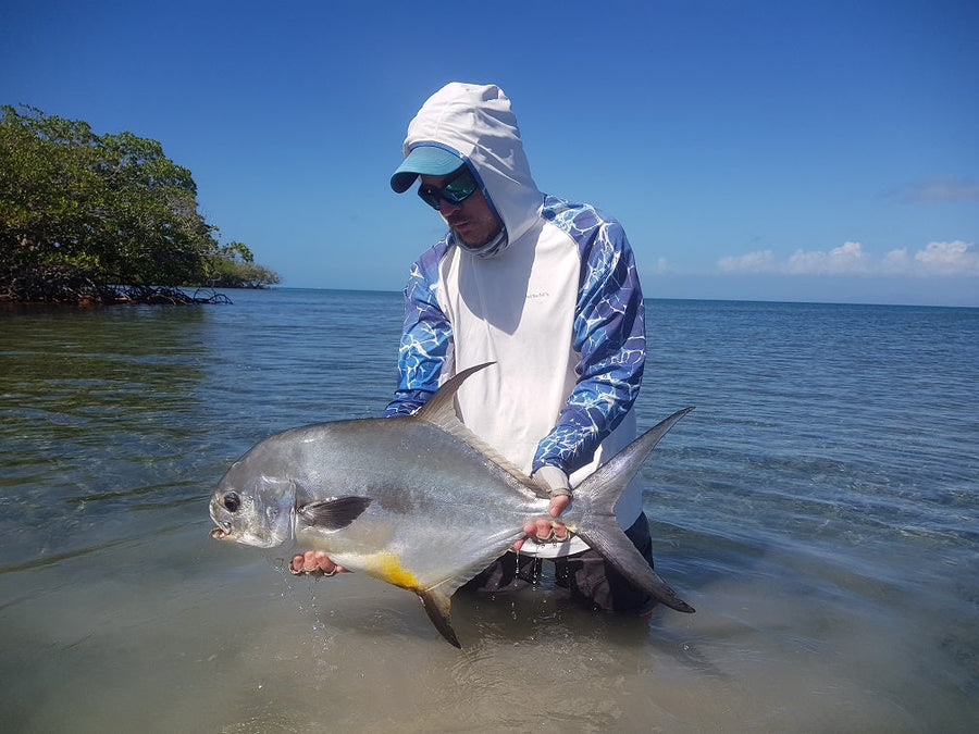 Fly angler holding a large permit in shallow Belize flats near mangroves under a clear blue sky