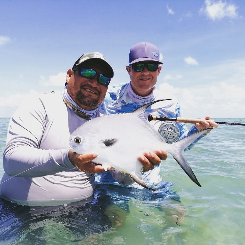 Two anglers holding a silver permit while wading during a Pesca Maya fly fishing trip in shallow clear water