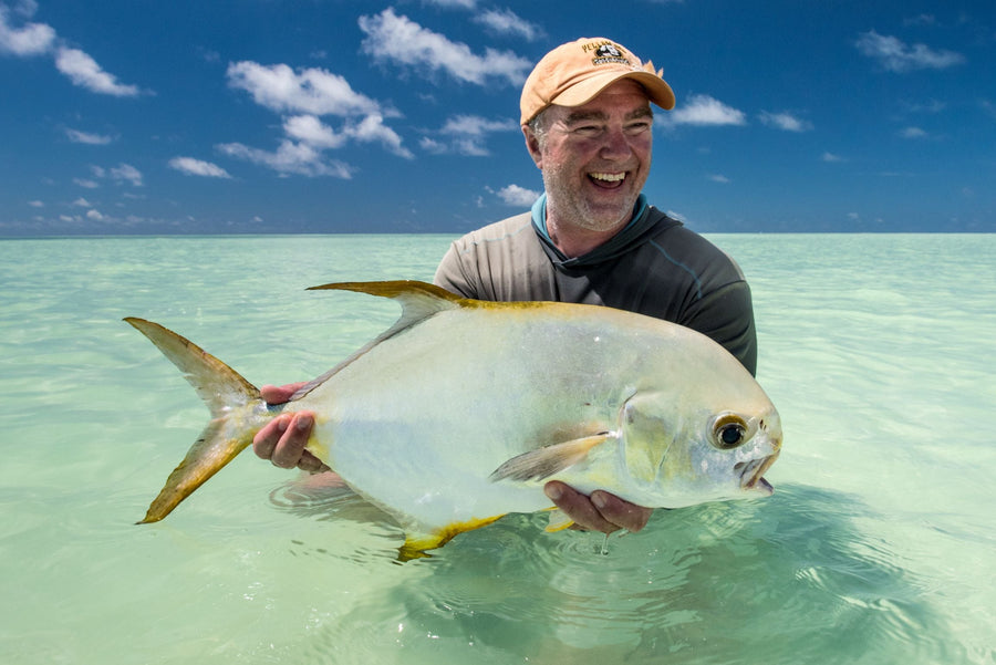 Smiling angler holding a large permit in shallow turquoise water on a sunny fly fishing trip