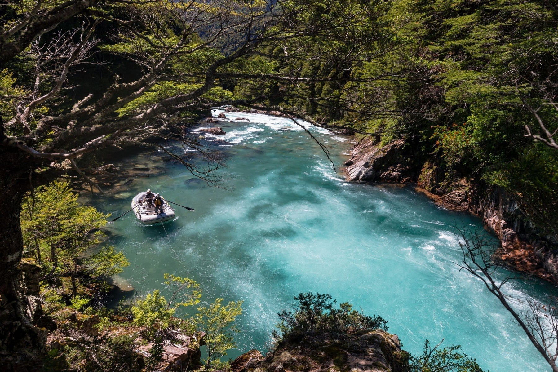 Two people navigate a turquoise river in an inflatable raft through a forested Patagonia landscape