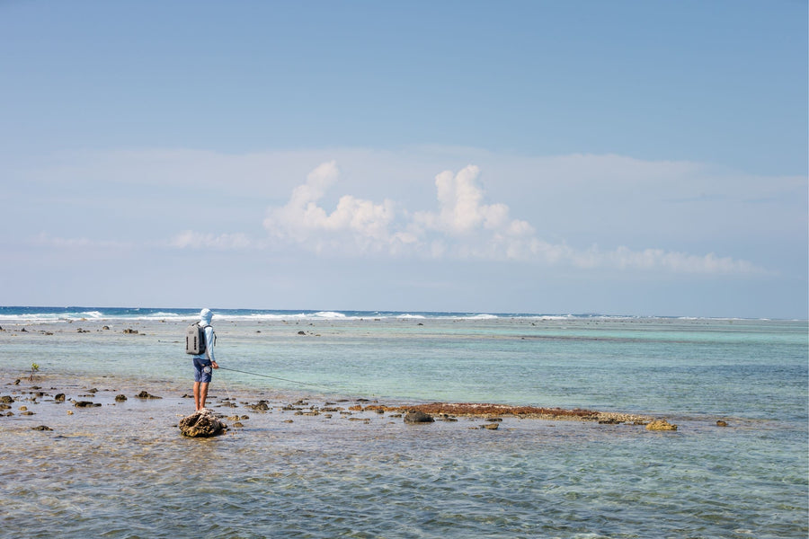 Angler fly fishing on shallow Belize flats, casting from a rocky reef into clear turquoise water under a blue sky