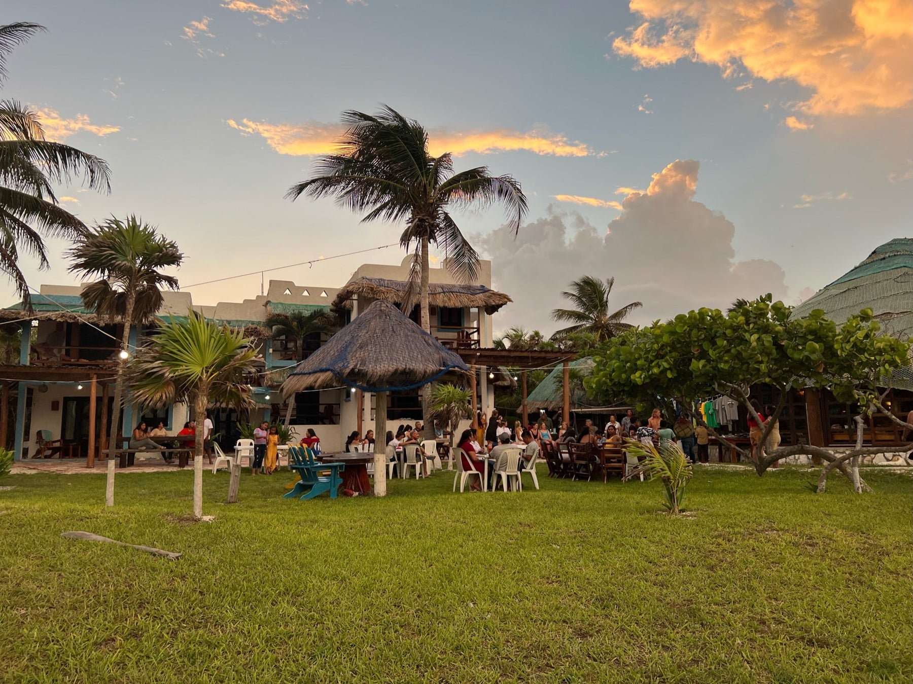 People gather outdoors at a tropical thatched-roof venue surrounded by palm trees at sunset