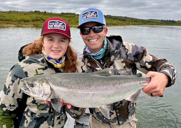 Two anglers on a fly-fishing trip holding a large silver salmon on a skiff in a river with grassy shoreline and overcast sky