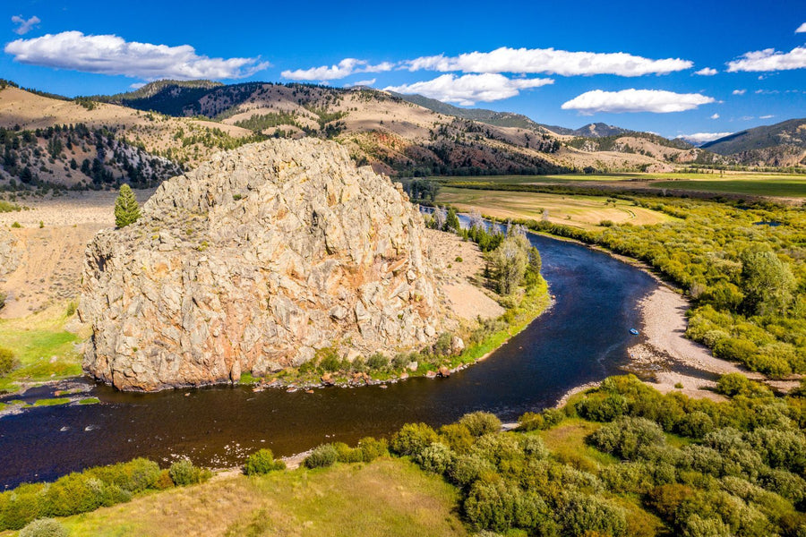 Aerial view of a winding river curving around a large rocky outcrop in a sunlit mountain valley with a small raft on the shore