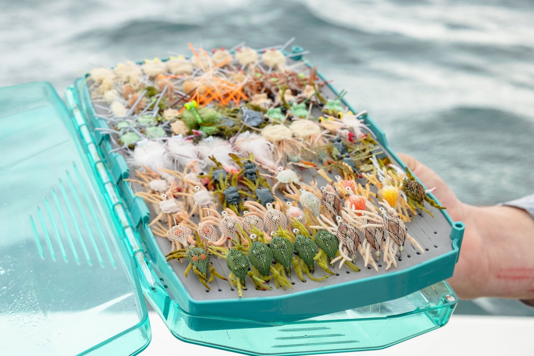 Top view of a fly box filled with colorful permit flies showcasing varied fly patterns with water in the background