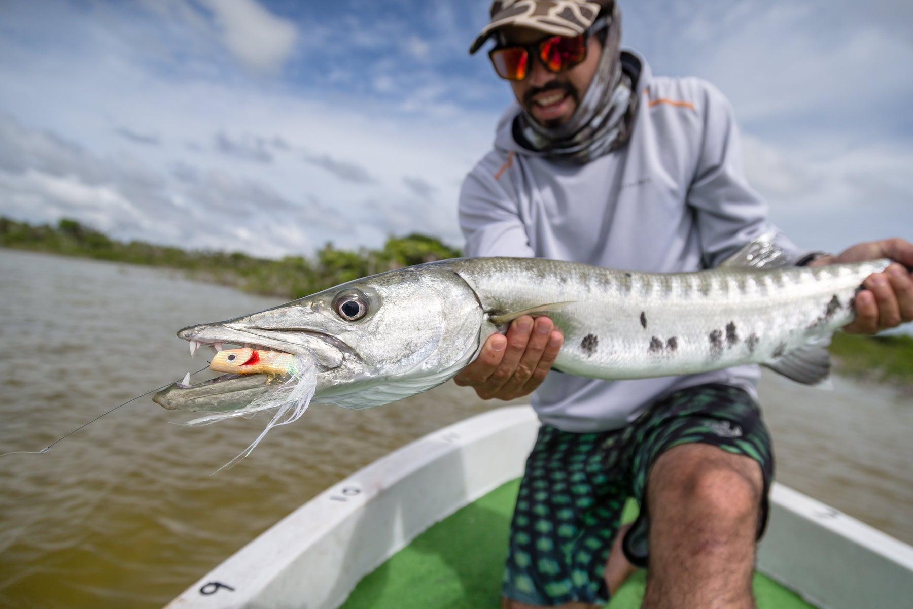 Angler holding a barracuda with a fly in its mouth aboard a boat in shallow coastal water