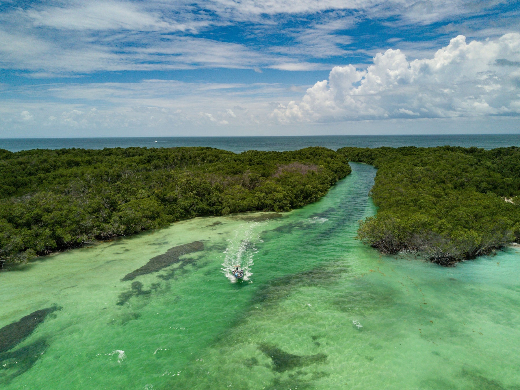 Aerial view of a small boat traveling through turquoise water between mangroves, with ocean and cloudy sky beyond