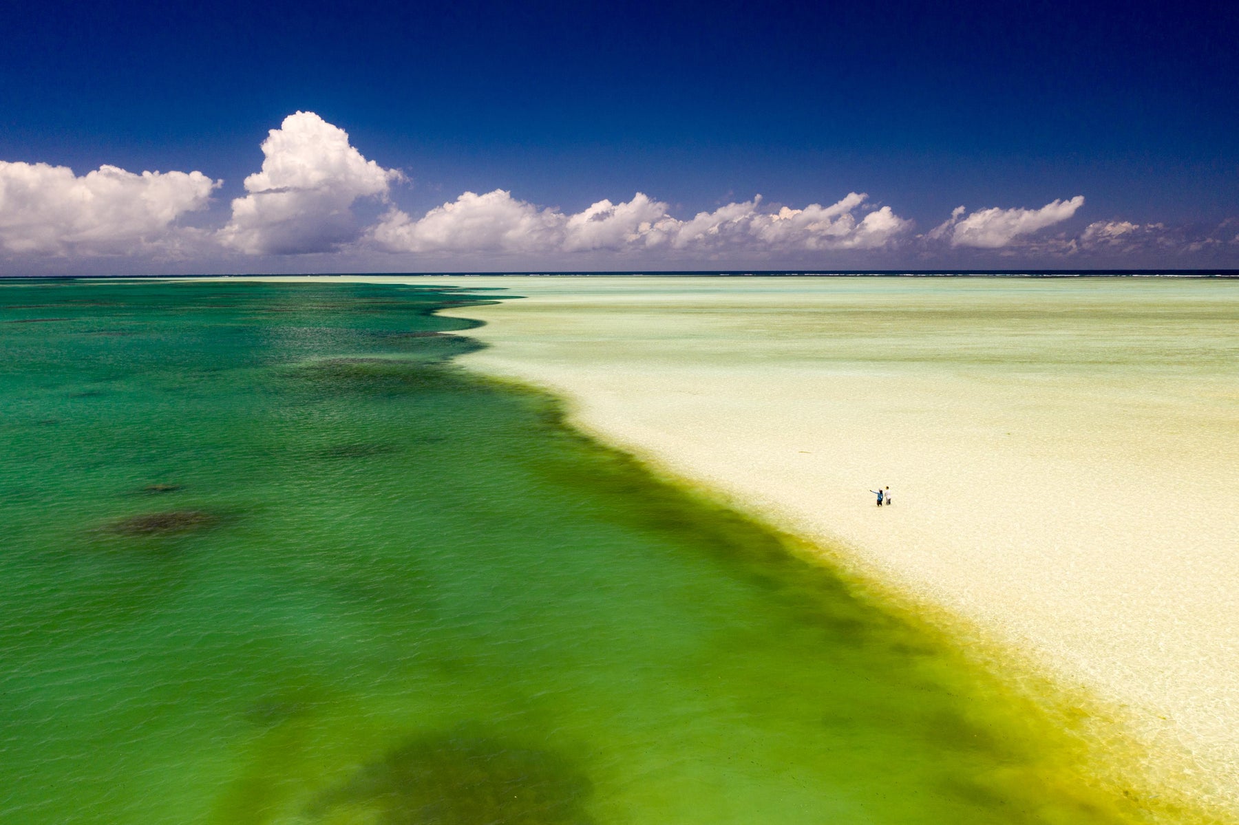 Two people stand on Alphonse atoll’s turquoise shallows and sand flats beneath towering clouds