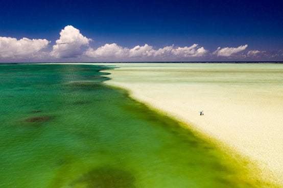 Two people stand on Alphonse atoll’s turquoise shallows and sand flats beneath towering clouds