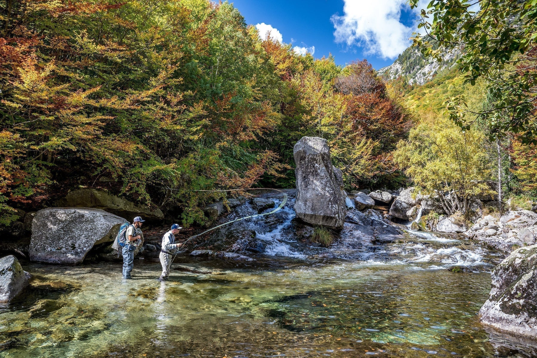 Two anglers fly fishing in a clear mountain stream surrounded by autumn forest and rocky peaks