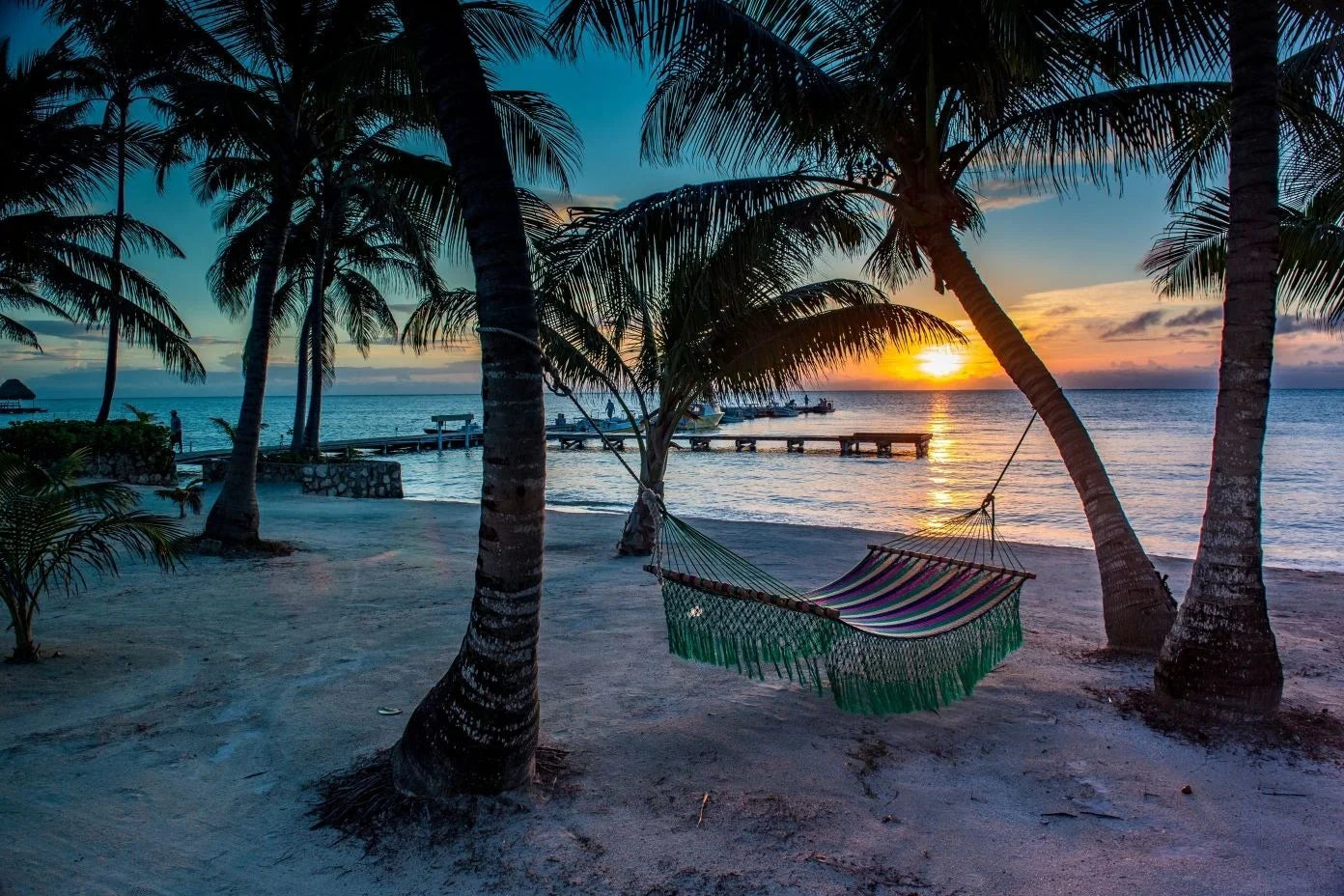 Hammock hanging between palm trees on a sandy beach at sunset with a dock and boats on the horizon