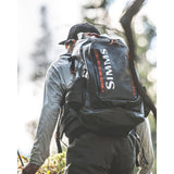 Rear view of Simms G3 Guide Backpack in Anvil showcasing roll top waterproof design and padded hipbelt on angler in forest