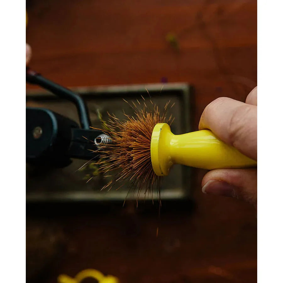 Close-up of Ergo Hair Packer for fly-tying, yellow ergonomic handle held against a hook in a vise showing dense brown hair bristles on a wooden bench