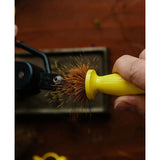 Close-up of Ergo Hair Packer for fly-tying, yellow ergonomic handle held against a hook in a vise showing dense brown hair bristles on a wooden bench